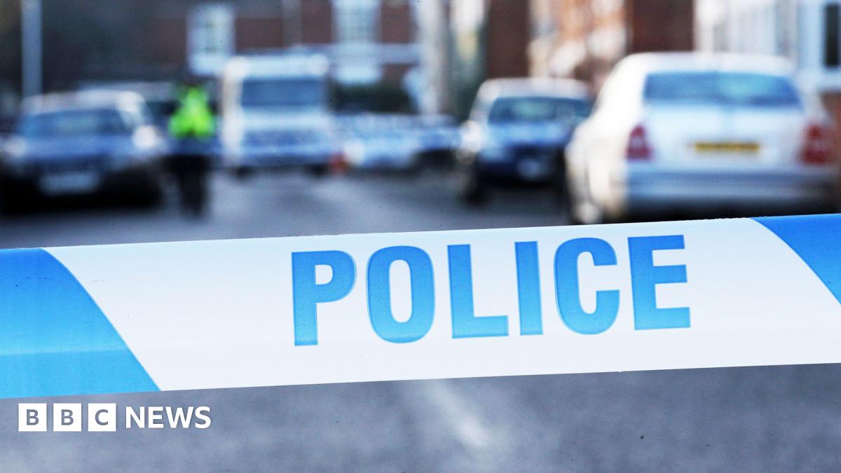A close up shot of blue and white police tape. It is striped with "police" written on it in blue capitals. In the background there are a number of cars on a residential street, although it is blurred quite heavily.