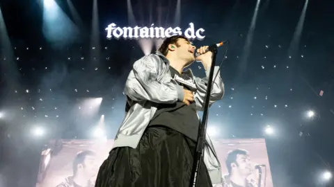 Shirlaine Forrest/Getty Images Grian Chatten of Fontaines DC, a man with short, dark hair, on stage at Wythenshawe Park on 15 August, 2025.  He is singing into a microphone and is wearing a silver sports jacket over a black t-shirt.  A similar image of him is also projected onto large screens behind him. The band's logo, in bright, lower case caligraphy font, is projected above his head. 