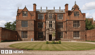 The grand building stands behind a circular lawn and flora. It has five sections with windows on three levels and an ornate frontage and entrance.