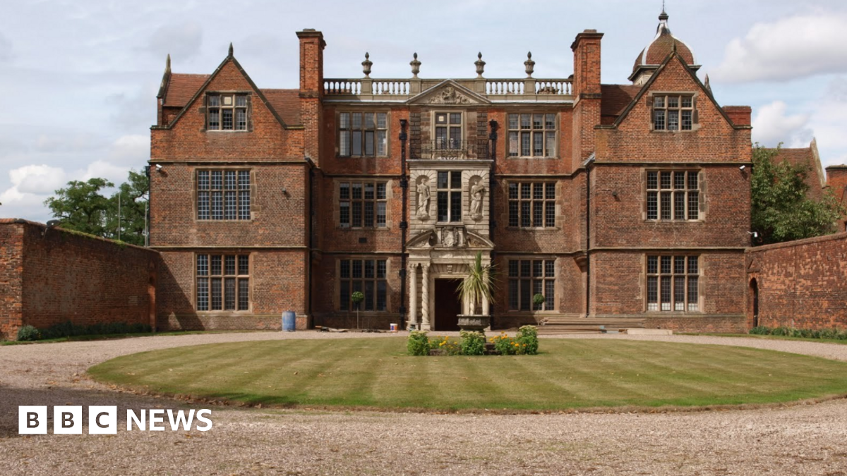The grand building stands behind a circular lawn and flora. It has five sections with windows on three levels and an ornate frontage and entrance.