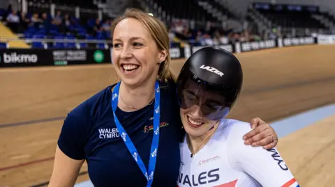 SWPIX Rachel Draper with her arm around a smiling Lowri Thomas who's in Welsh cycling kit and wearing a track helmet. They're next to a cycling track. Rachel has her blonde hair tied back and is wearing a blue lanyard and navy t-shirt. 