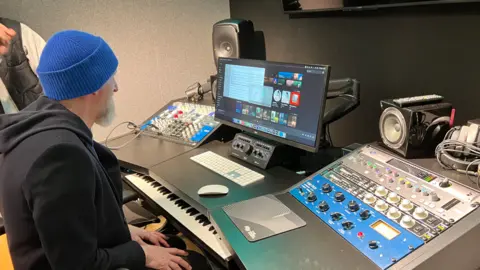 Ian Dench sitting at a mixing desk with mixers and a computer screen with a keyboard under the desk, just visible. 
Ian is wearing a black hoody and a blue beanie hat. There are a number of speakers on the desk and soft studio walls.