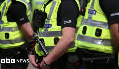 Three Police Scotland officers wearing hi-vis vests over black clothing with "Police Scotland" in white writing on the upper arm.