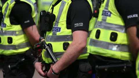 PA Media Three Police Scotland officers wearing hi-vis vests over black clothing with "Police Scotland" in white writing on the upper arm.