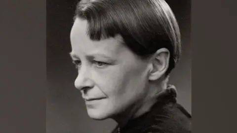 Ramsay & Muspratt A black and white photo of Nan Youngman. She is in profile and slightly turning to the right so both her eyes can be seen. She has a relaxed face and short sleek hair, tucked behind her ear. She is wearing a dark top. 