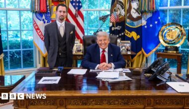Trump sits behind desk in blue suit and red tie while Antoni stands in three-piece gray suit with a beige tie, white collar and watch chain in front of windows covered in colorful flags and banners
