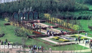 People walk round flower beds on part of the exhibition site. Pathways and colourful flower beds can be seen across a green lawn.