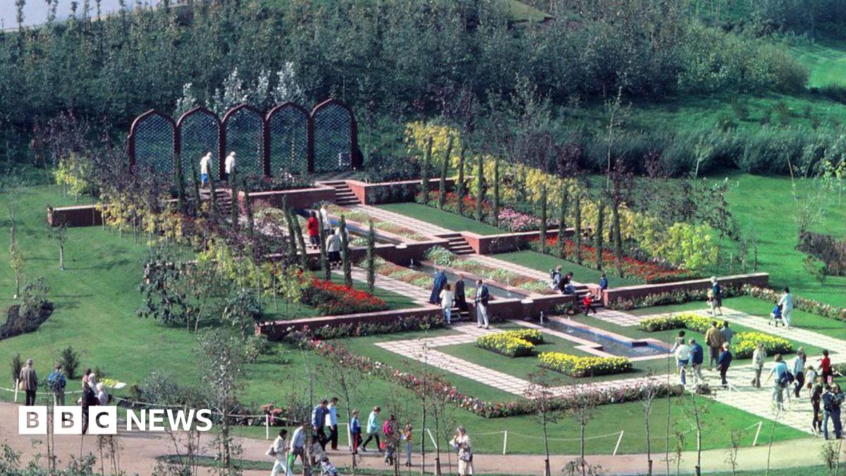 People walk round flower beds on part of the exhibition site. Pathways and colourful flower beds can be seen across a green lawn.