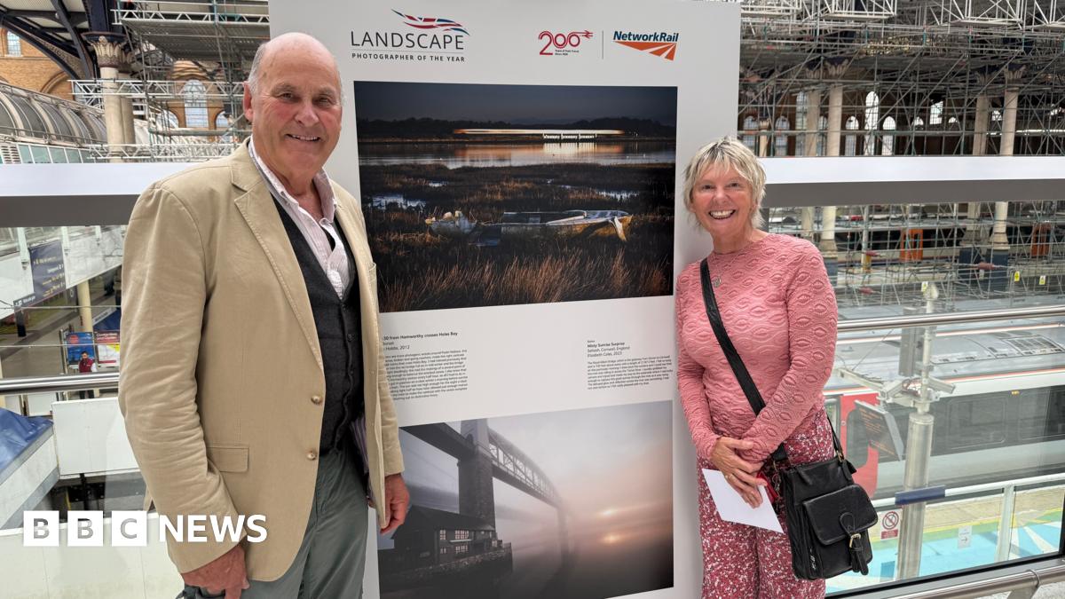 Charlie Waite, wearing a shirt, waistcoat and jacket stands in front of a photo on display. Also next to the photo is Elizabeth Coles wearing a pink jumper and pink patterned trousers.