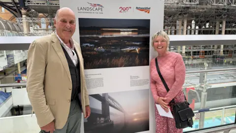 Network Rail Charlie Waite, wearing a shirt, waistcoat and jacket stands in front of a photo on display. Also next to the photo is Elizabeth Coles wearing a pink jumper and pink patterned trousers.