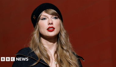 Taylor Swift smiling wearing a black beanie hat and black jacket.