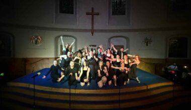 Large cast photo pose with arms out, black background, all seen in the spotlight. Church background shown.