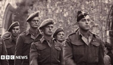 A black and white image showing men from the Cambridgeshire Regiment marching past Ely's war memorial in late 1945. They are in Army uniform. They are being watched by men, women and children.