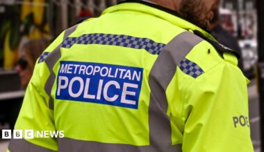 A police officer wearing a high-visibility jacket with the words "Metropolitan Police" on the back, standing outdoors.