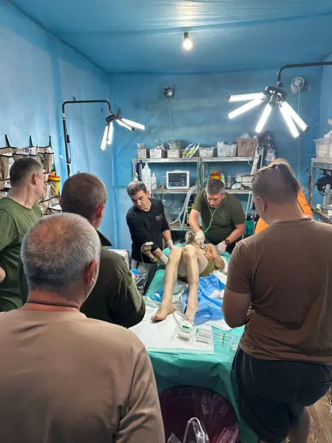 A man wearing just shorts lie on a bed, surrounded by six other men. There are shelves on the walls with various medical items, and medical items also rest on the bed next to the man.