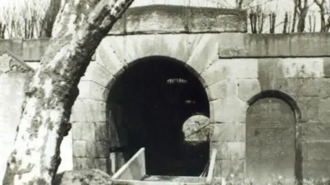 Birmingham City Council Archives A black and white stone archway that opens up a dark tunnel. There is a grave to the right of it and numerous trees in front of it and behind it. 