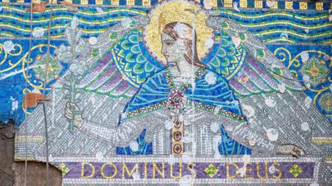 Great Places Housing Group A mosaic frieze consisting of decorative Venetian glass tiles in white, purple, gold and blue, depicting an angel with the Latin 'Dominus Deus' running a long a banner underneath. The display is placed uncovered on a partially demolished wall.