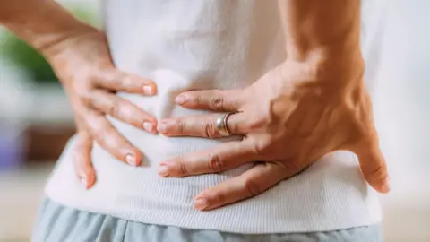 Getty Images A person is facing away from the camera with both hands clutching their back. They are wearing a white top and pale blue trousers. 