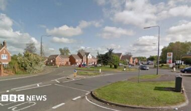A road with houses along the left hand side and a garage up and on the right. There is a roundabout with a silver car going round it and grass verges with trees in the background.