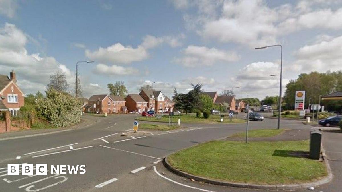 A road with houses along the left hand side and a garage up and on the right. There is a roundabout with a silver car going round it and grass verges with trees in the background.