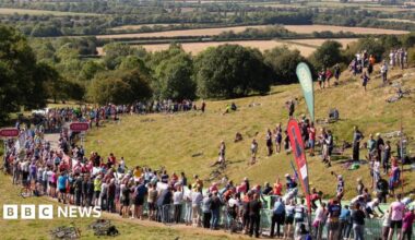 A road at the foot of a grassy hill is lined with spectators.