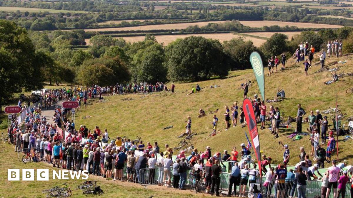 A road at the foot of a grassy hill is lined with spectators.