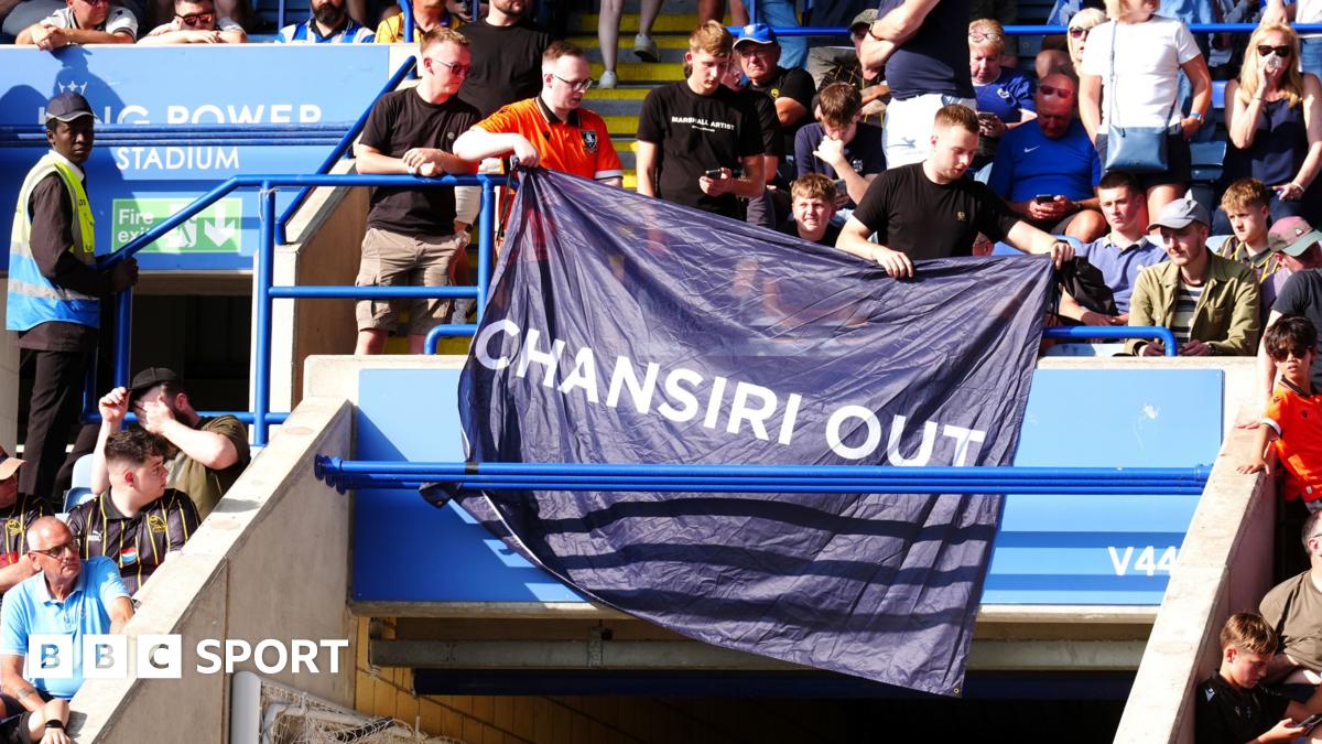 Black banner that says 'SWFC for sale - enough is enough' at the back of a stadium with red seats where numerous people sit