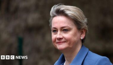 A close up of Yvette Cooper as she walks past some bushes. She is smiling slightly and wearing a blue jacket and white leaf-shaped earrings. Her hair is cropped short.
