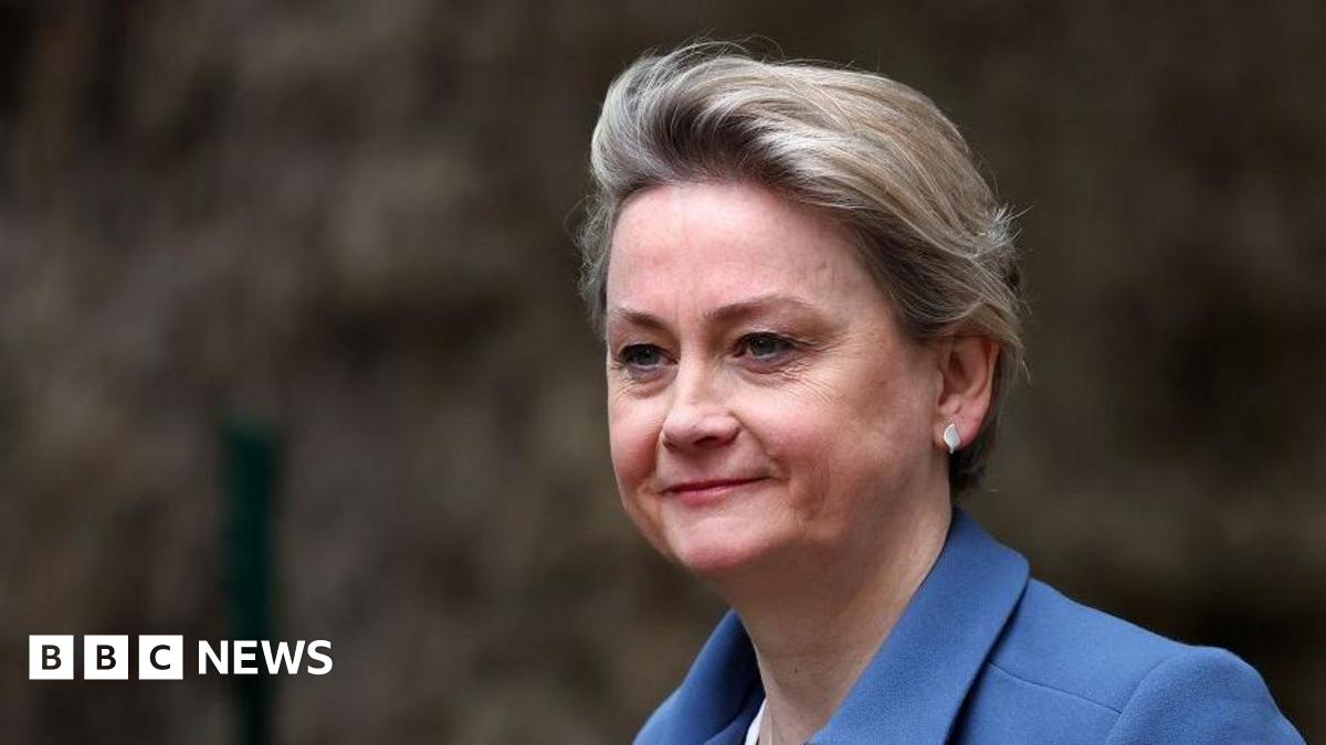 A close up of Yvette Cooper as she walks past some bushes. She is smiling slightly and wearing a blue jacket and white leaf-shaped earrings. Her hair is cropped short.