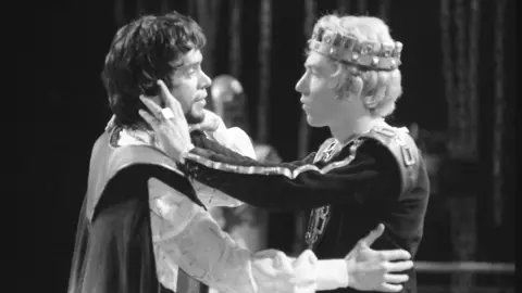 A black and white shot of two men in period costume in a production of Shakespeare's Edward II - one wears a king's crown. They are locked in a distant embrace, the king with his hand's on the other man's face. 