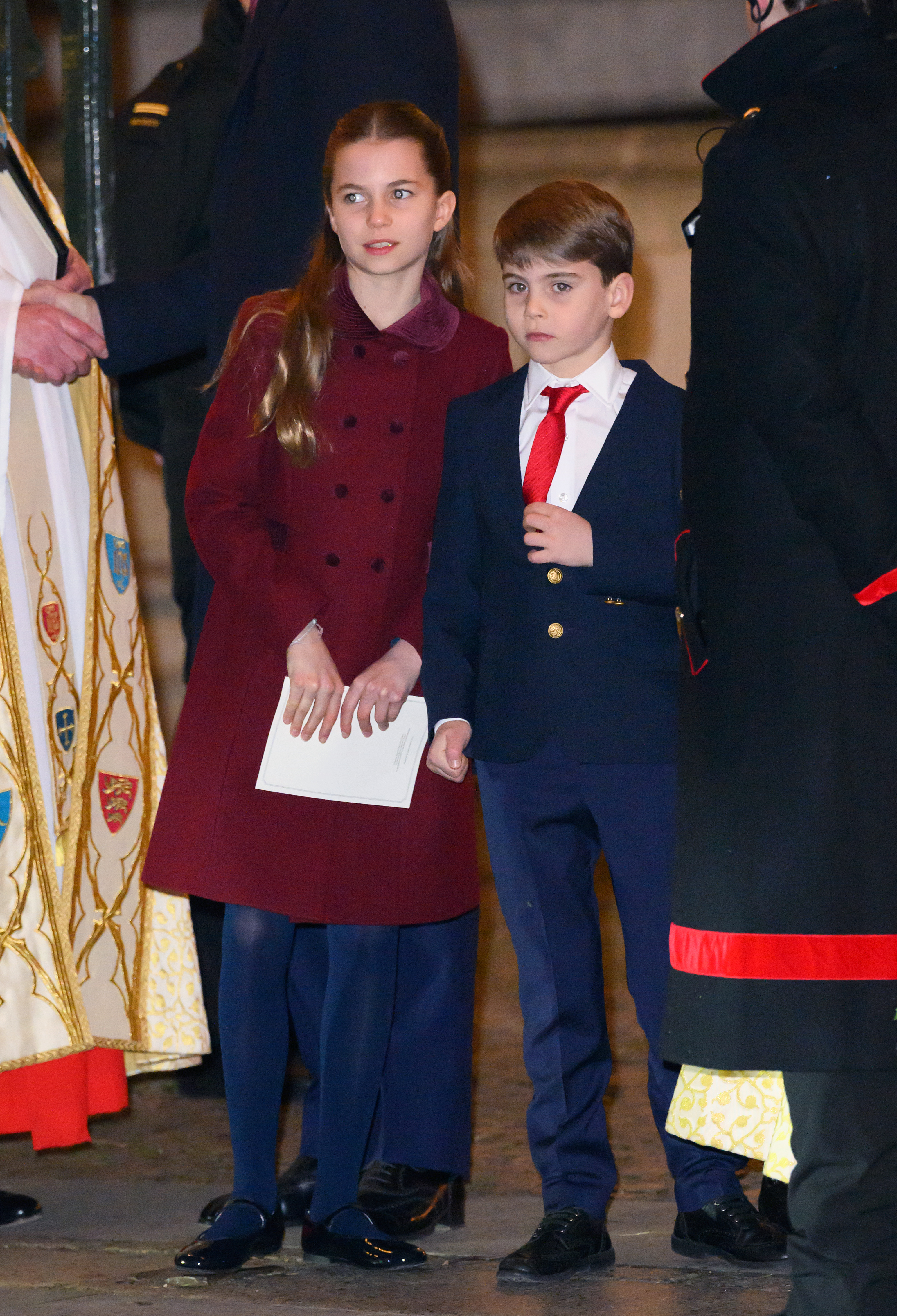 Prince Louis wearing a blue suit and Princess Charlotte wearing a maroon coat standing in Westminster Abbey