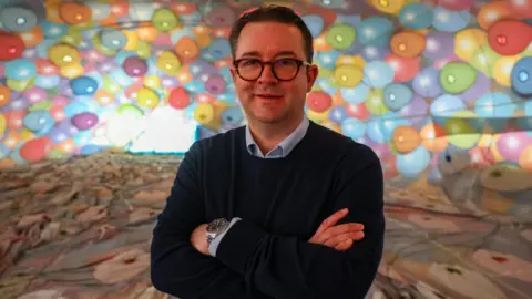Paul Gillis Ben Hardy. He has short dark hair and is wearing glasses with thick black frames. He is standing in front of a hot air balloon envelope which is multi coloured. Ben is wearing a blue collared shirt and a navy blue jumper. He is standing with his arms crossed, looking at the camera and smiling. 