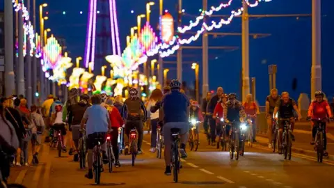 Visit Blackpool Blackpool prom at dusk with dozens of cyclists seen at a distance riding under Blackpool Illuminations