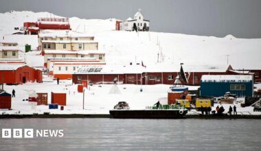 A series of single storey building coloured brown and orange stand in the snow comprising Chile's Presidente Eduardo Frei military base next to the Antarctic Ocean. People can be seen working along the shoreline and there is an open-topped boat in the water.