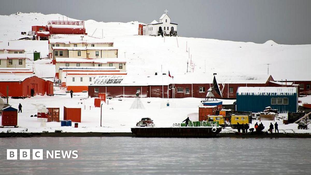 A series of single storey building coloured brown and orange stand in the snow comprising Chile's Presidente Eduardo Frei military base next to the Antarctic Ocean. People can be seen working along the shoreline and there is an open-topped boat in the water.