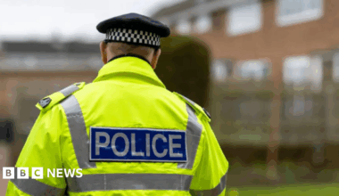 A stock image showing a male police officer wearing a chequered black and white cap and a yellow hi-vis jacket which says 'POLICE' across the back in reflective material. He is facing away from the camera towards a brick building with a tall hedge outside.