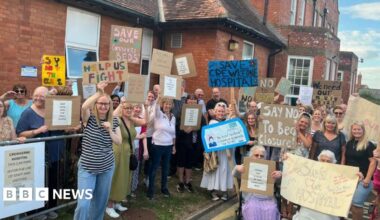 A crowd of people stand outside a red brick building carrying signs saying "Save Crewkerne Hospital", "say no to bed closure" and "help us fight".