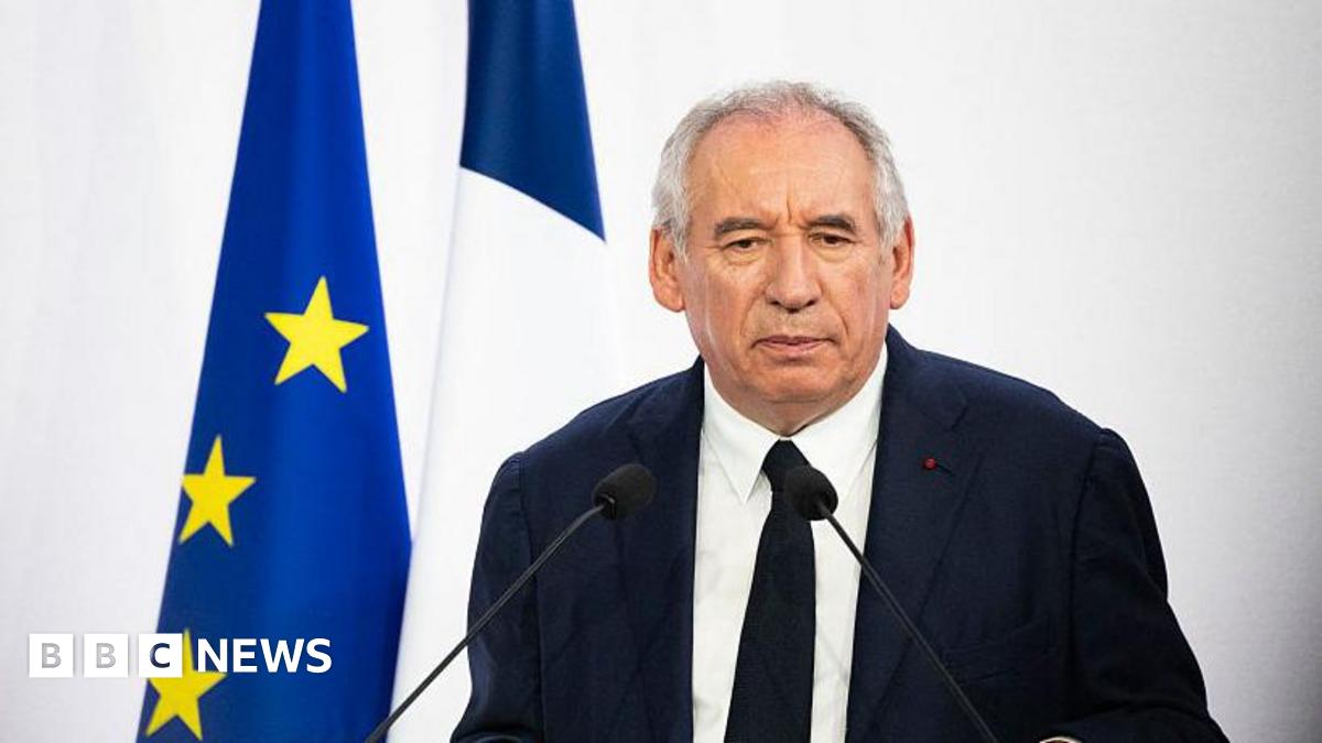 The French prime minister wears a dark jacket and tie and stands in front of a French and European Union flag