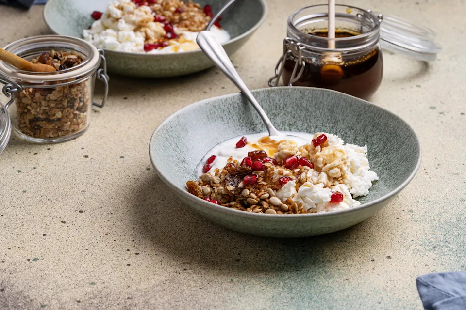 Two bowls of yogurt, granola, cottage cheese and dried fruit sit on a beige countertop.