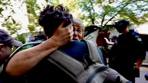 BBC News A blonde woman, who looks distressed, tightly embraces a man with short brown hair.  Her hand is round the back of his head and only the back of his head, back and rucksack can be seen.
