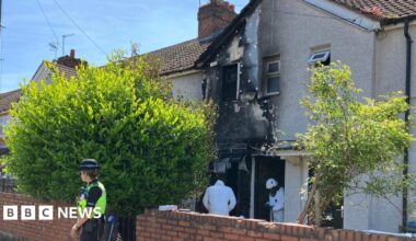 A terraced two-storey house shows fire damage on the ground floor, first floor outer wall, window and part of the roof. Two people in a forensic outfit are at the front door while a policewoman is guarding the property outside.