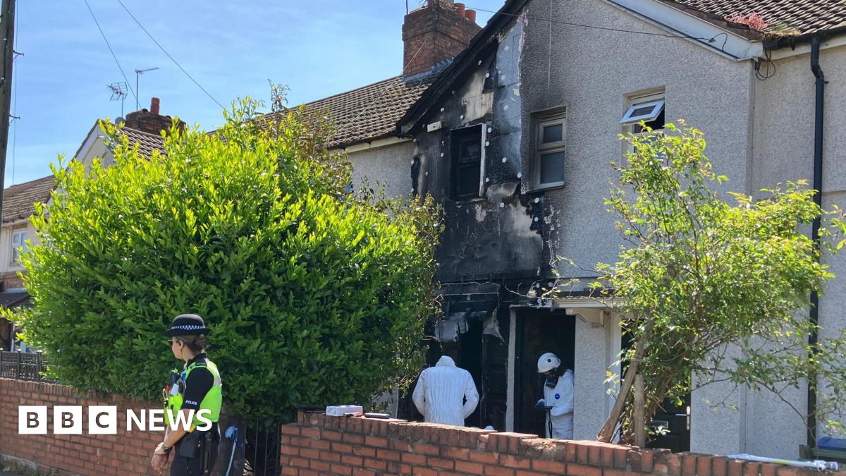 A terraced two-storey house shows fire damage on the ground floor, first floor outer wall, window and part of the roof. Two people in a forensic outfit are at the front door while a policewoman is guarding the property outside.