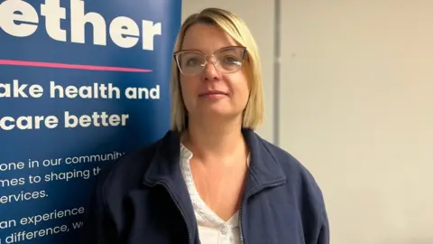 BBC A woman standing in front of a blue promotional board with blonde hair and is wearing glasses with a white shirt and blue fleece zip-up top.