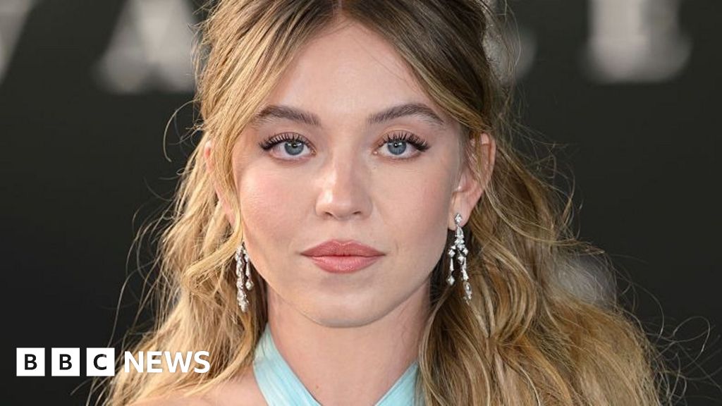 Sydney Sweeney poses with long wavy brown hair, wearing silver earrings and a light blue dress (not the American Eagle advert)