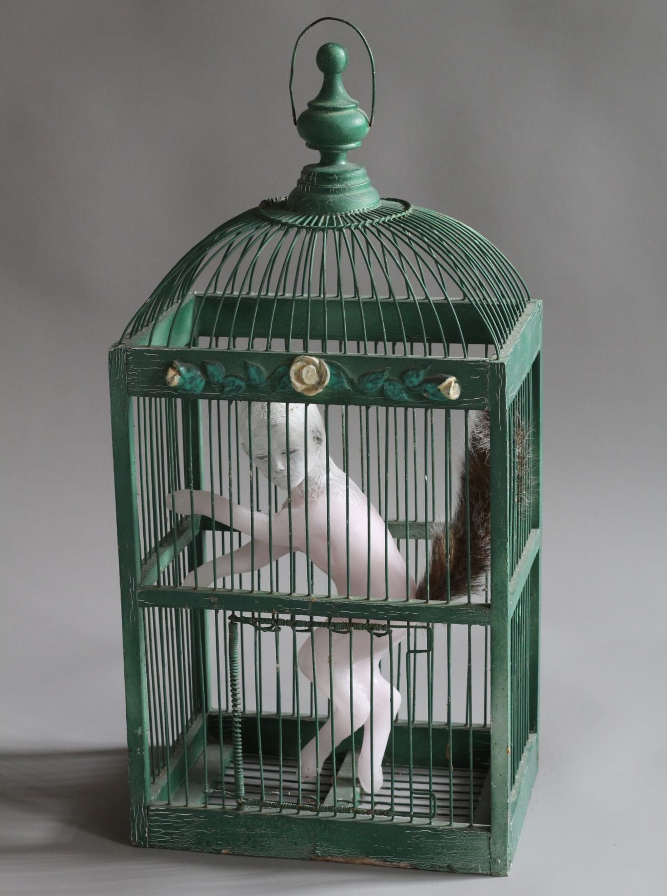 a green bird cage with a glass and ceramic sculpture of a child with a fox's tail inside