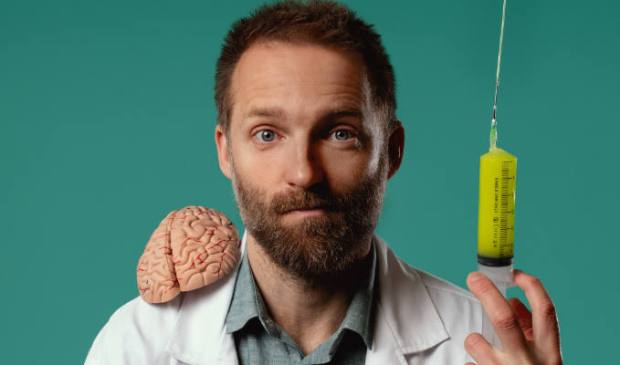 A man in a lab coat stands against a teal background, holding a large syringe in one hand and displaying a brain on his shoulder.