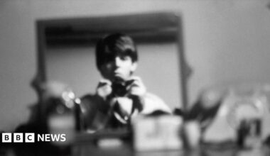 Titled: George, Ringo, and John backstage at The Beatles Christmas Show, Finsbury Park Astoria, London, December 1963. The three bandmates are sat down and look down the lens of the camera. They wear dark suits and are captured in black and white film.