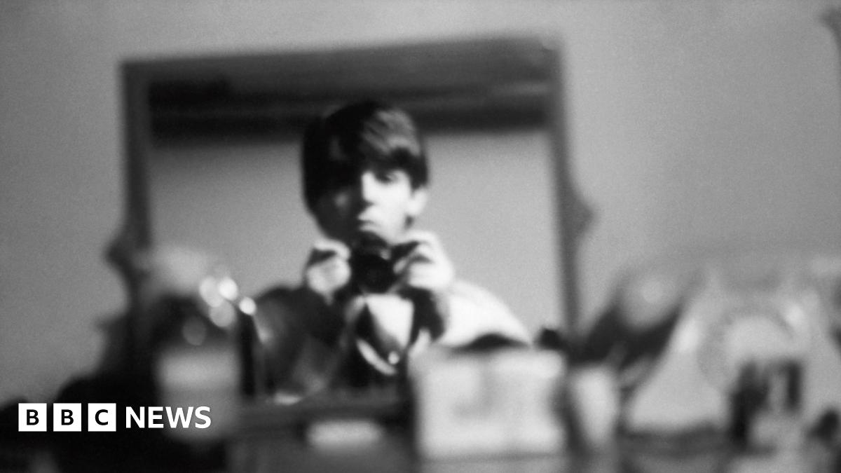 Titled: George, Ringo, and John backstage at The Beatles Christmas Show, Finsbury Park Astoria, London, December 1963. The three bandmates are sat down and look down the lens of the camera. They wear dark suits and are captured in black and white film.