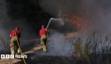 Two firefighters with a yellow hose pipe are directing water on to a grass fire. They are wearing red tops and light coloured trousers and facing the flames.