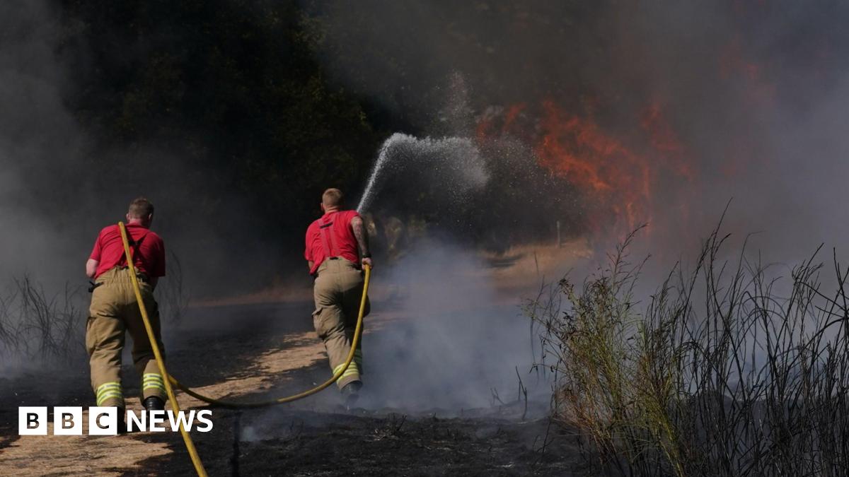 Two firefighters with a yellow hose pipe are directing water on to a grass fire. They are wearing red tops and light coloured trousers and facing the flames.